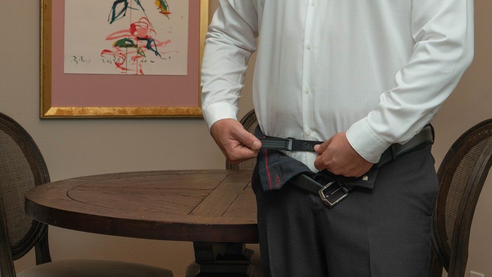 A man in a white dress shirt fastening the Stuck Tuck belt over his shirt, below his open trousers